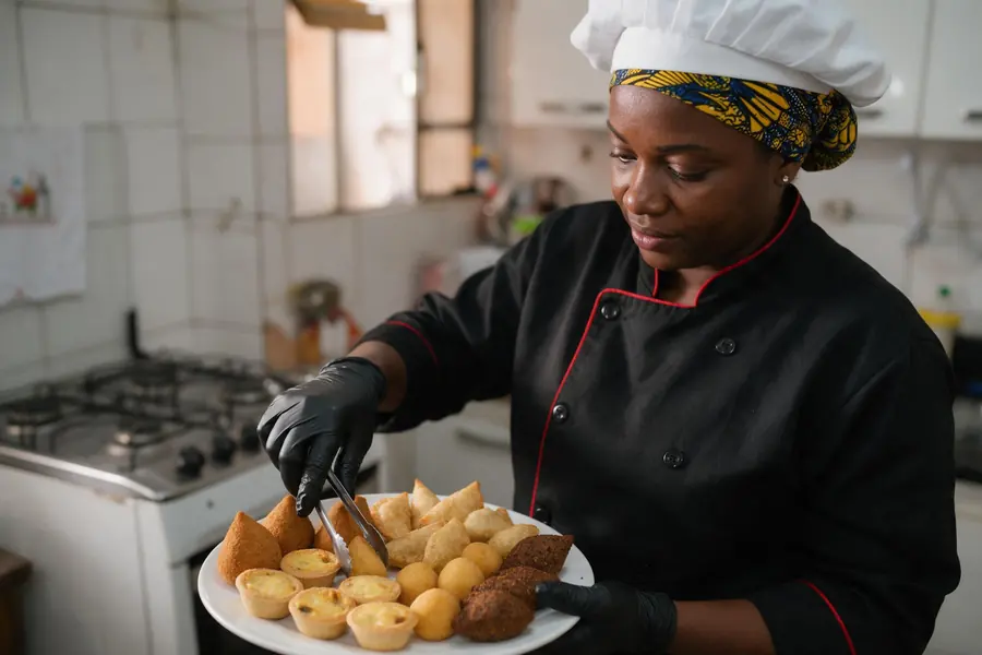 Cozinheira negra de uniforme arruma coxinhas, empadinhas e pastelzinhos numa travessa com pegador e luvas pretas